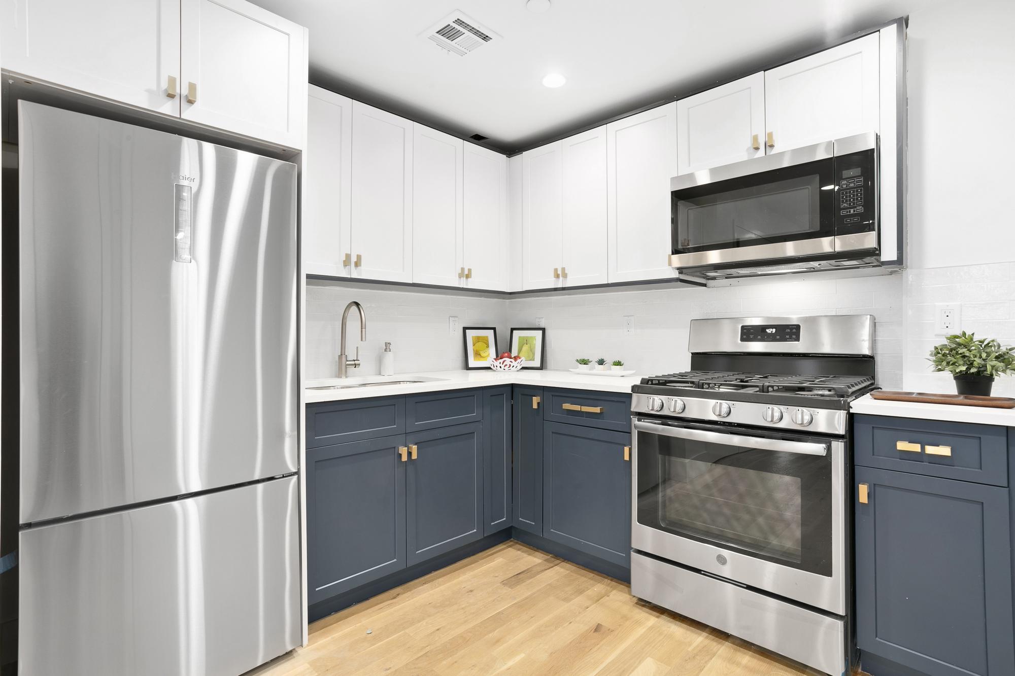 Modern Kitchen with White Top Cabinetry, Dark Slate Blue Lower Cabinetry with Wood Handles, Stainless Steel Fridge, Oven and Microwave, White Backsplash, Light Wood Flooring, Chrome Sink Faucet
